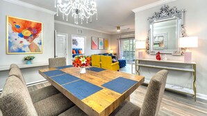 A dining area with a wooden table, blue placemats, and a chandelier. There are chairs around the table, and a sofa with yellow cushions in the background. - Splendid Renovated 2 + 2 Condo Near Disney with Amazing Amenities (Davenport)