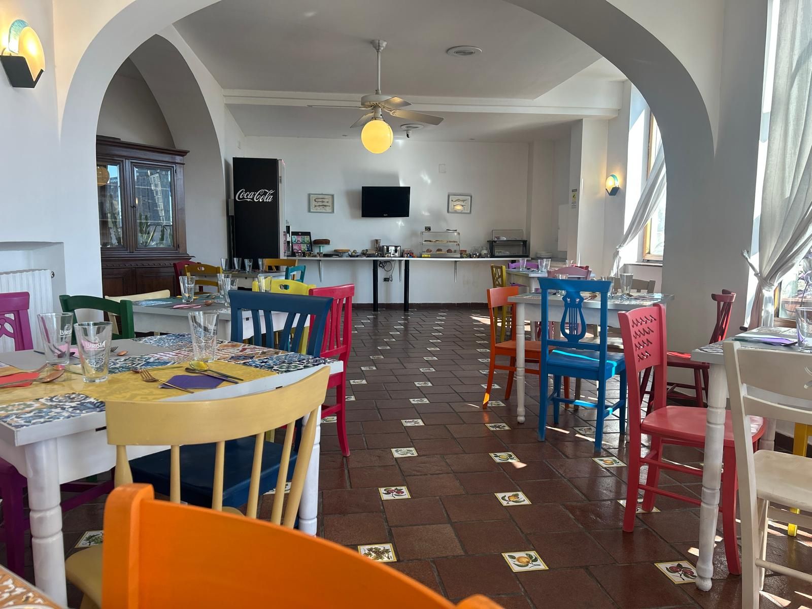 Ein Restaurant mit mehreren Tischen und Stühlen, einem Deckenventilator und einem Coca-Cola-Kühlschrank.