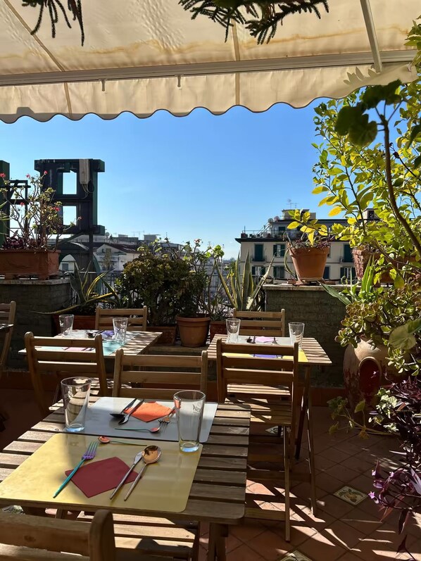 An outdoor patio with wooden tables and chairs, potted plants, and a clear view of the cityscape in the background.