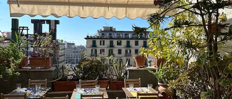An outdoor dining area with tables and chairs, potted plants, and a view of buildings in the background.