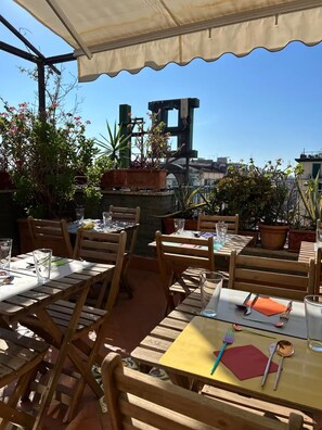 Un patio exterior con mesas y sillas de madera, plantas en macetas y una vista despejada del paisaje urbano.