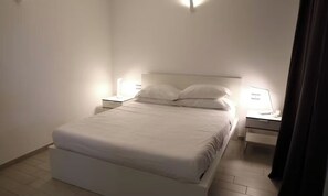 A neatly made bed in a hotel room with a white color scheme, a bedside table with a lamp, and a nightstand with a lamp.