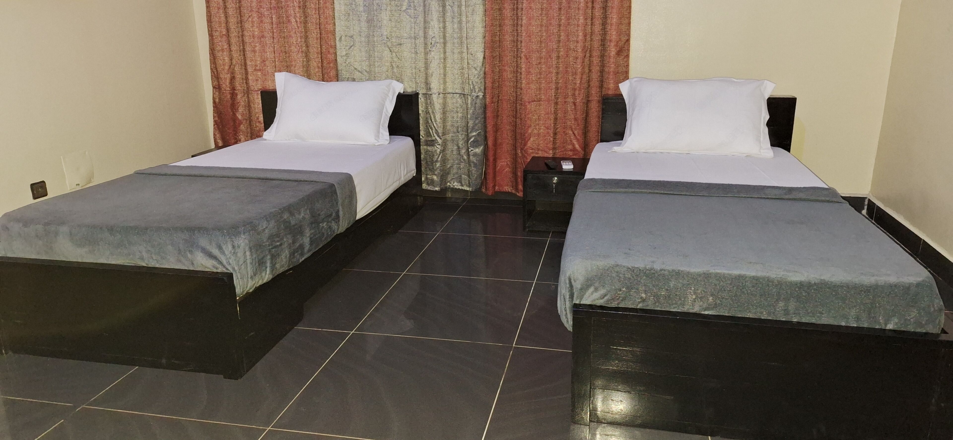 A room with two single beds, each with a grey bedspread and white pillows, a small bedside table, and a curtain with a red and beige pattern.
