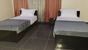 A room with two single beds, each with a grey bedspread and white pillows, a small bedside table, and a curtain with a red and beige pattern.