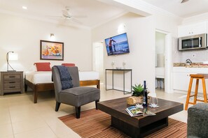 A modern living room with a bed, a gray armchair, a coffee table with magazines, a TV, and a kitchen area with a microwave. - Charming Studio With Garden Oasis in Kingston (Kingston)