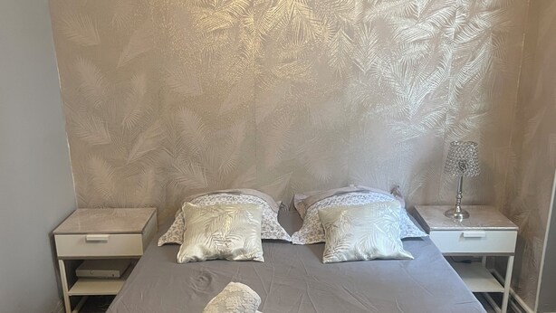 Ein ordentlich bezogenes Bett mit einer braunen Tagesdecke und zwei Kissen, ein kleiner Nachttisch mit einer Lampe und eine Wand mit Blättermotiv.