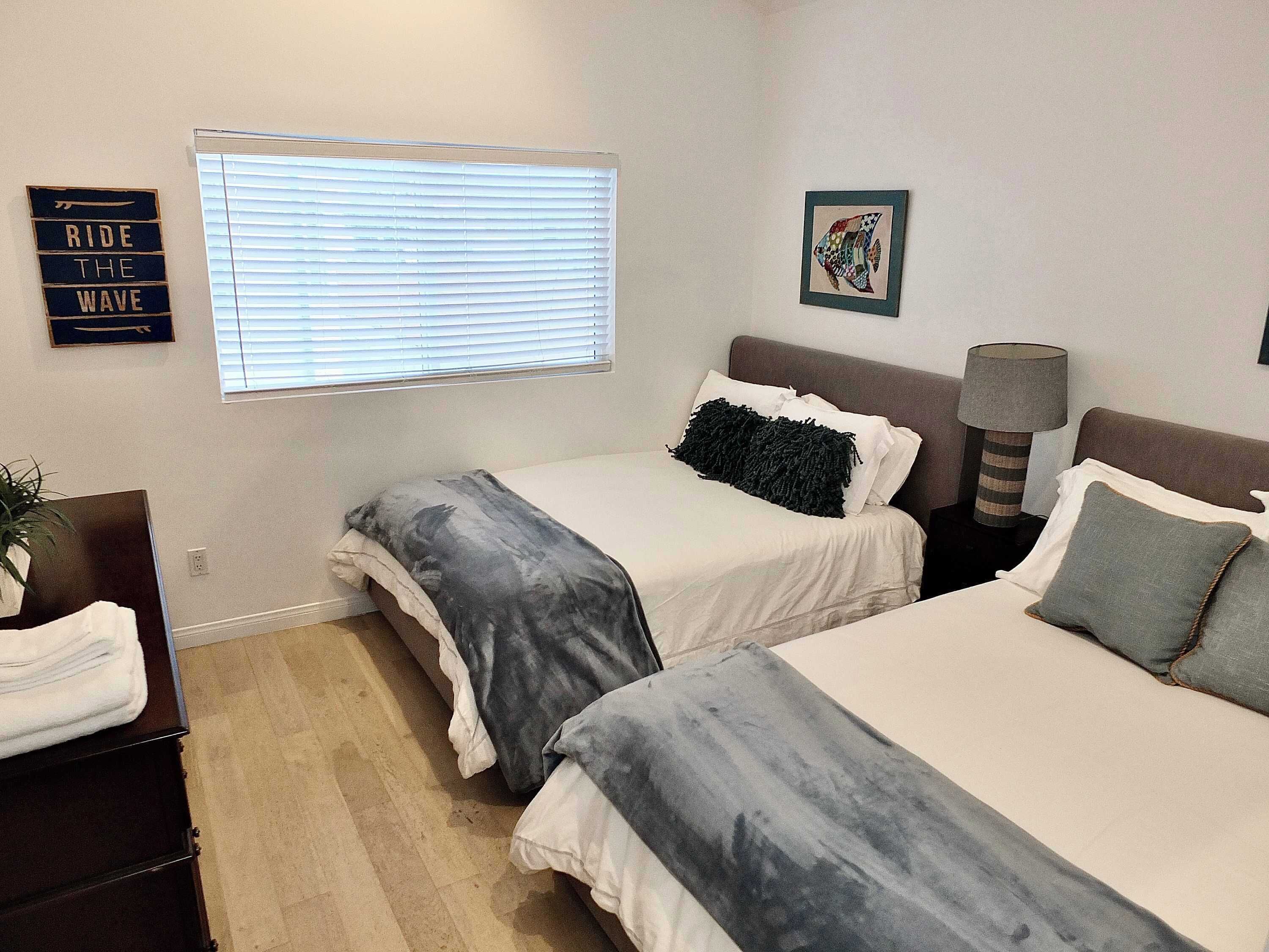 A bedroom with two beds, a bedside lamp, a wall-mounted sign, and a window with blinds.