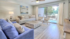 A living room with a sofa, coffee table, and chairs, a painting on the wall, and a sliding glass door leading to an outdoor area.