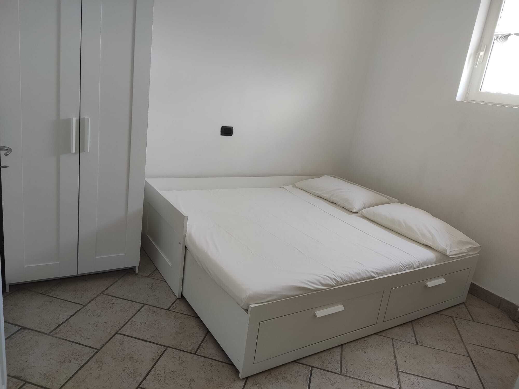 Una habitació petita i blanca amb un llit, un armari i una finestra.