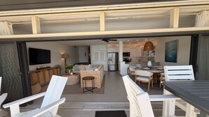 Un espacio de estar al aire libre con techo, con sillas y una mesa blancos, una cocina con muebles blancos y un comedor con mesa y sillas.