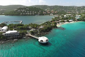 Vista aérea de una zona costera con un edificio en forma de cúpula cerca del agua, un muelle y una playa con edificios al fondo.