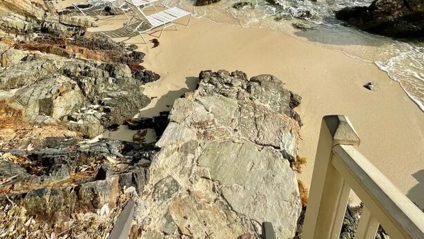 Una playa con agua turquesa cristalina, costa rocosa y un barco a lo lejos.