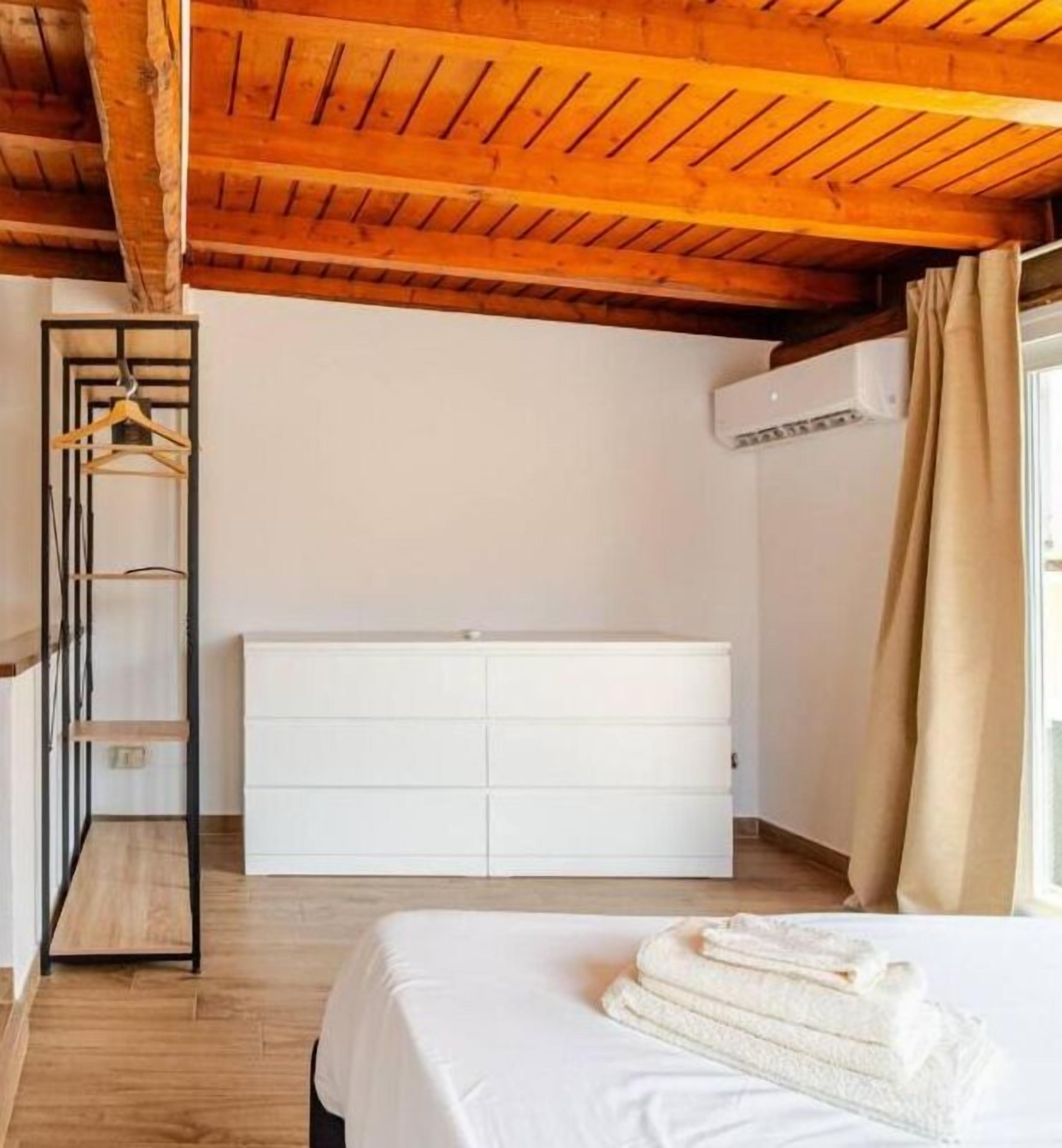 A bedroom with a wooden ceiling, a white dresser, a bed with white linens, and a wooden shelf unit.