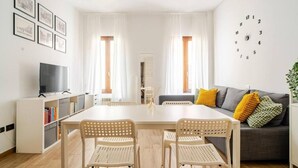 A modern living room with a dining table, two chairs, a sofa with cushions, a bookshelf, a television, and a wall clock. - La Terrazza su Brescia · Panoramic terrace (Brescia)