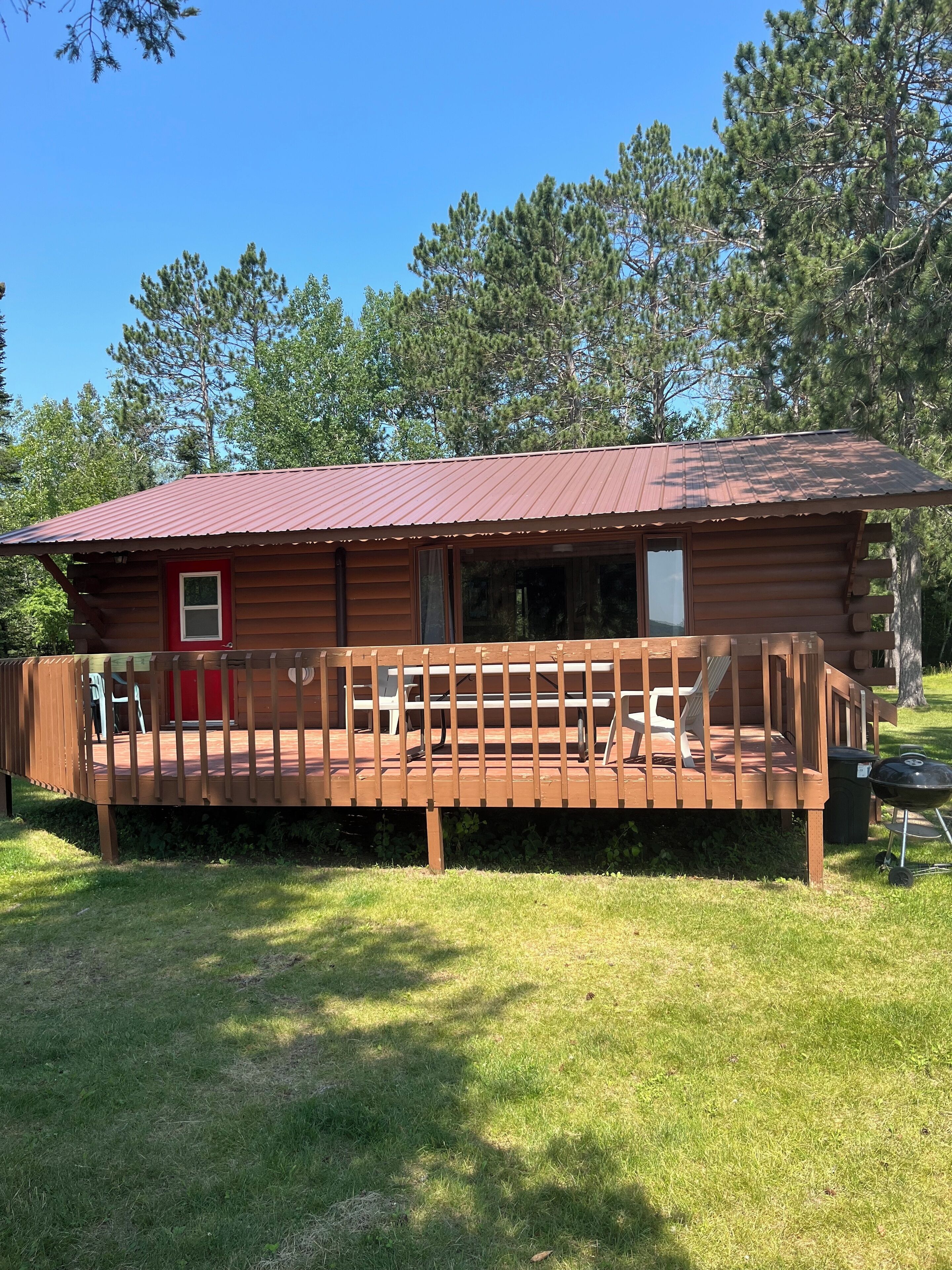 Une cabane en bois avec une porte rouge, une véranda dotée d’une balustrade et un barbecue à l’extérieur.