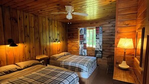Een kamer in chaletstijl met twee bedden, een plafondventilator en een raam met uitzicht op bomen.