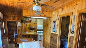Een gezellig interieur van een vakantiehuisje, met houten plafond, plafondventilator, keukenhoek en een woonkamer met een bank en een fauteuil met patroon.