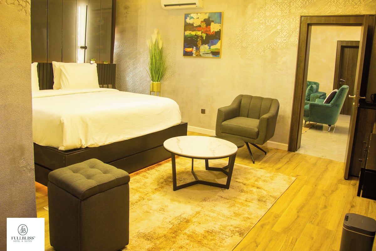 A modern hotel room with a large bed, a small round table, a green armchair, and a potted plant.