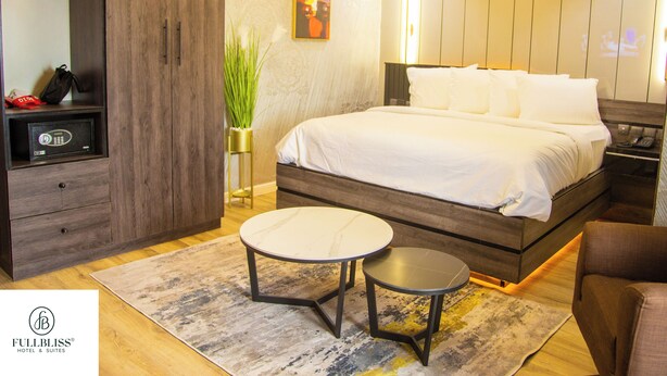 Une chambre d’hôtel moderne avec un lit, une table de chevet, une chaise et une petite table ronde. On y trouve également un coffre-fort, une plante en pot et un tableau accroché au mur.