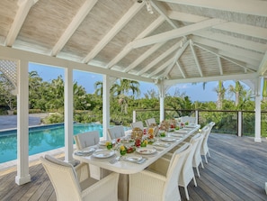 Un área de comedor exterior techada con vista a la piscina, mobiliario de ratán blanco y una mesa dispuesta para cuatro personas.