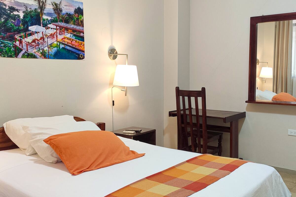 Habitación de hotel con cama, escritorio, silla, lámpara de pared, un cuadro y espejo.