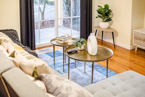 Un salon avec un canapé, une table basse, une petite table d’appoint avec une plante et une fenêtre avec des rideaux.