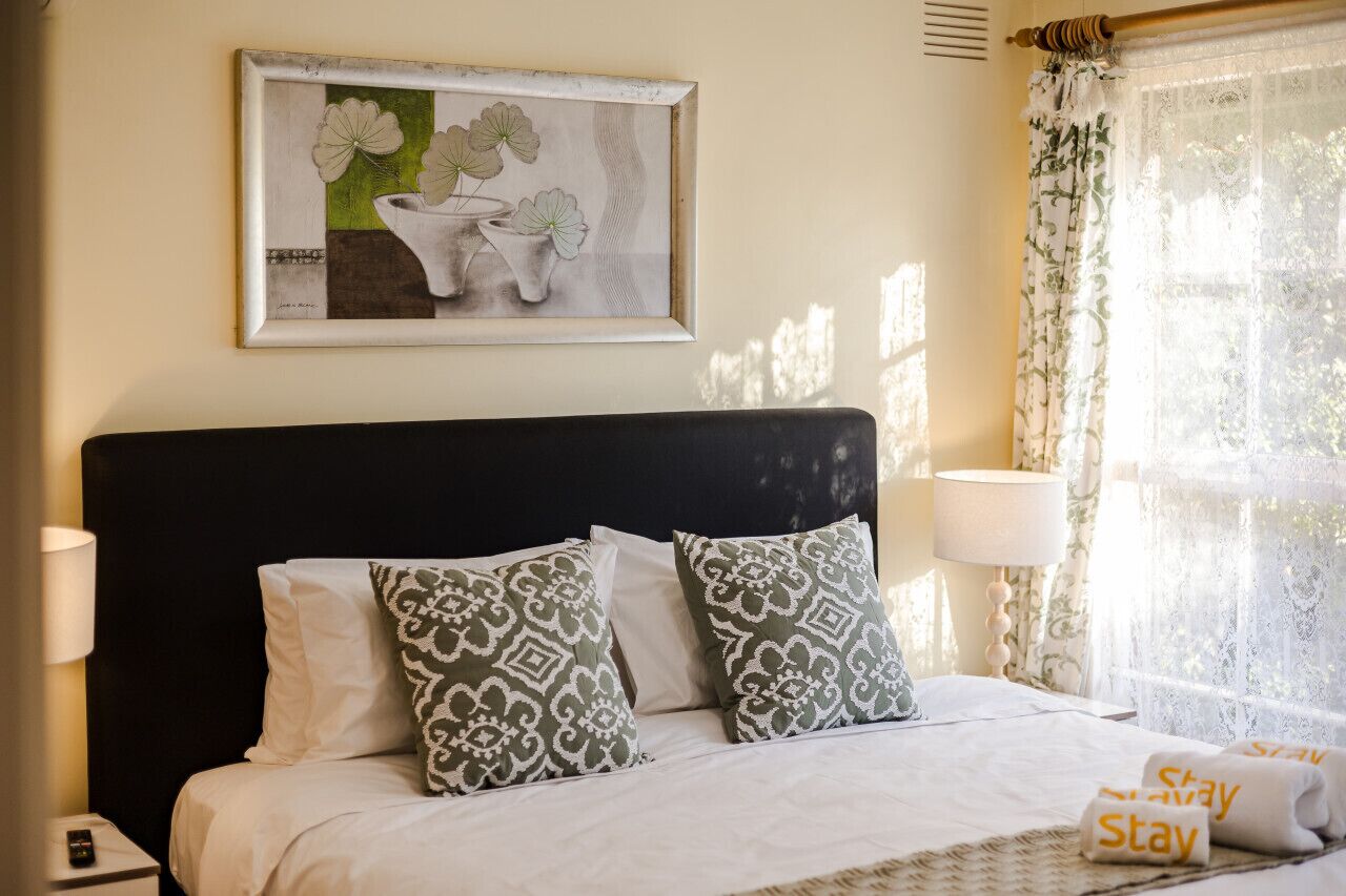 Una camera d'albergo con un letto, cuscini, un quadro, un comodino, una lampada e una finestra con tende.