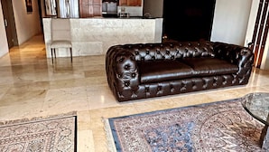 Sala de estar com sofá de couro marrom escuro, tapete estampado e área de cozinha com armários de madeira e ilha integrada.