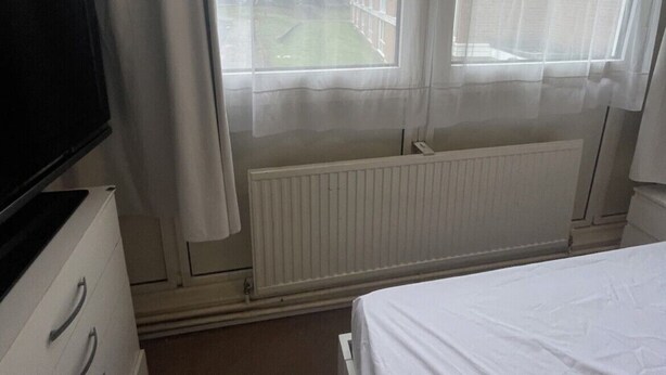 A hotel room with a bed, a television, a radiator, and a view of a building and greenery outside. - Lovely Double Bedroom - Putney (London)