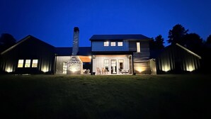 A modern two-story house with a large chimney, a covered patio with outdoor seating, and a well-lit front entrance. - The Reserve at Kirby Landing (Kirby)