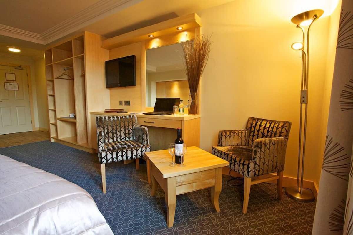 A hotel room with a bed, a desk, two patterned chairs, a small table with a bottle and glasses, a TV on the wall, and a floor lamp.