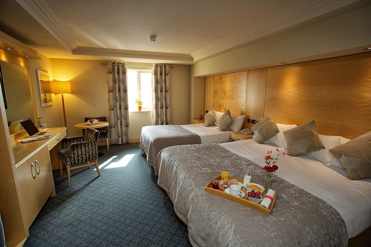 A hotel room with two beds, a desk with a laptop, a tray with breakfast items, and a window with curtains.