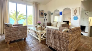 A spacious living area with wicker furniture, a glass coffee table, and a view of palm trees through large windows. - Villa Gardelina (Corsica)