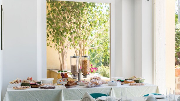 Un espace repas avec des tables préparées pour le petit-déjeuner, où l’on propose une variété de viennoiseries et de boissons, et d’où l’on peut admirer la vue sur les arbres et un pot de fleurs suspendu.