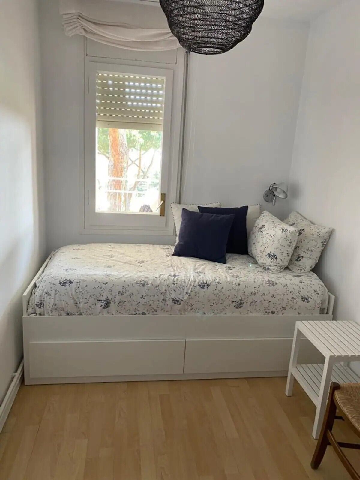 Un dormitorio pequeño y ordenado con una cama individual, una ventana con persianas, una lámpara colgante y una mesita.