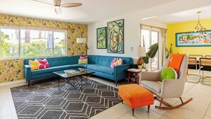 A living room with a teal sofa, orange ottoman, and a rocking chair. There are two paintings on the wall, a ceiling fan, and a window with a view of greenery.