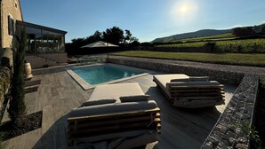 A swimming pool with lounge chairs and a pergola, surrounded by a stone wall and a view of a vineyard.