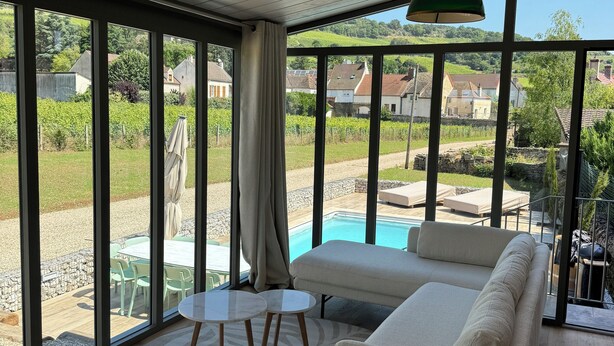 A modern living room with a large window, a green pendant light, and a view of a swimming pool and greenery outside.