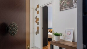 Un corridoio con una porta in legno, una parete bianca con specchi esagonali, un letto con testiera blu e un cartello di benvenuto.