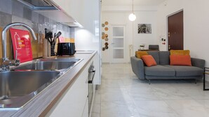 Una cucina moderna con lavello in acciaio inox, una macchina da caffè nera e un cartello informativo rosso. Nell'area soggiorno, un divano grigio con cuscini arancioni e una porta bianca con decorazione parietale geometrica.