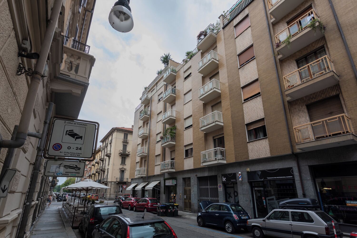 Una stretta via fiancheggiata da edifici a più piani, con balconi adornati da piante, un'area di parcheggio e un segnale che indica divieto di sosta.