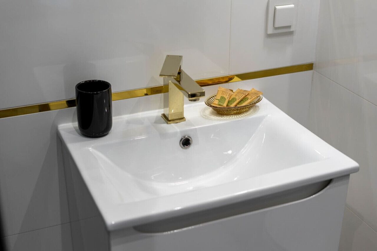 A white bathroom sink with a gold faucet, a black mug, and a bowl of toiletries.
