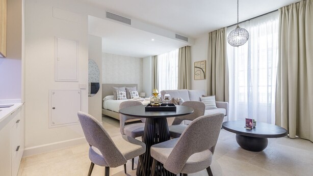 A modern hotel room with a dining area, a sofa, and a large window with curtains.