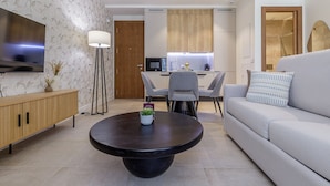 A modern living room with a flat-screen TV, a round coffee table, a grey sofa, and a dining area with a wooden cabinet and a lamp.