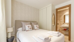 Habitación de hotel moderna con una cama grande, una mesita de noche con lámpara, un baño con lavamanos y espejo, y una puerta de madera.