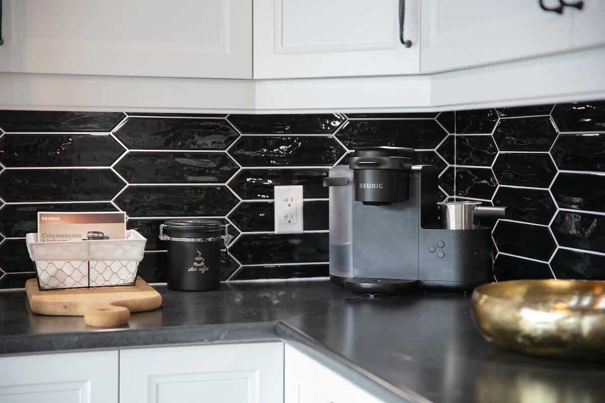 Une cuisine avec un dosseret de crédence en carreaux hexagonaux noirs et blancs, une machine à café Keurig, une cafetière et une planche à découper en bois, accompagnée d’une boîte de dosettes de café Keurig.