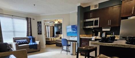 A modern kitchen with dark cabinets, a microwave, and a coffee maker. There is a dining area with a table and chairs, and a living room with a sofa and a TV.