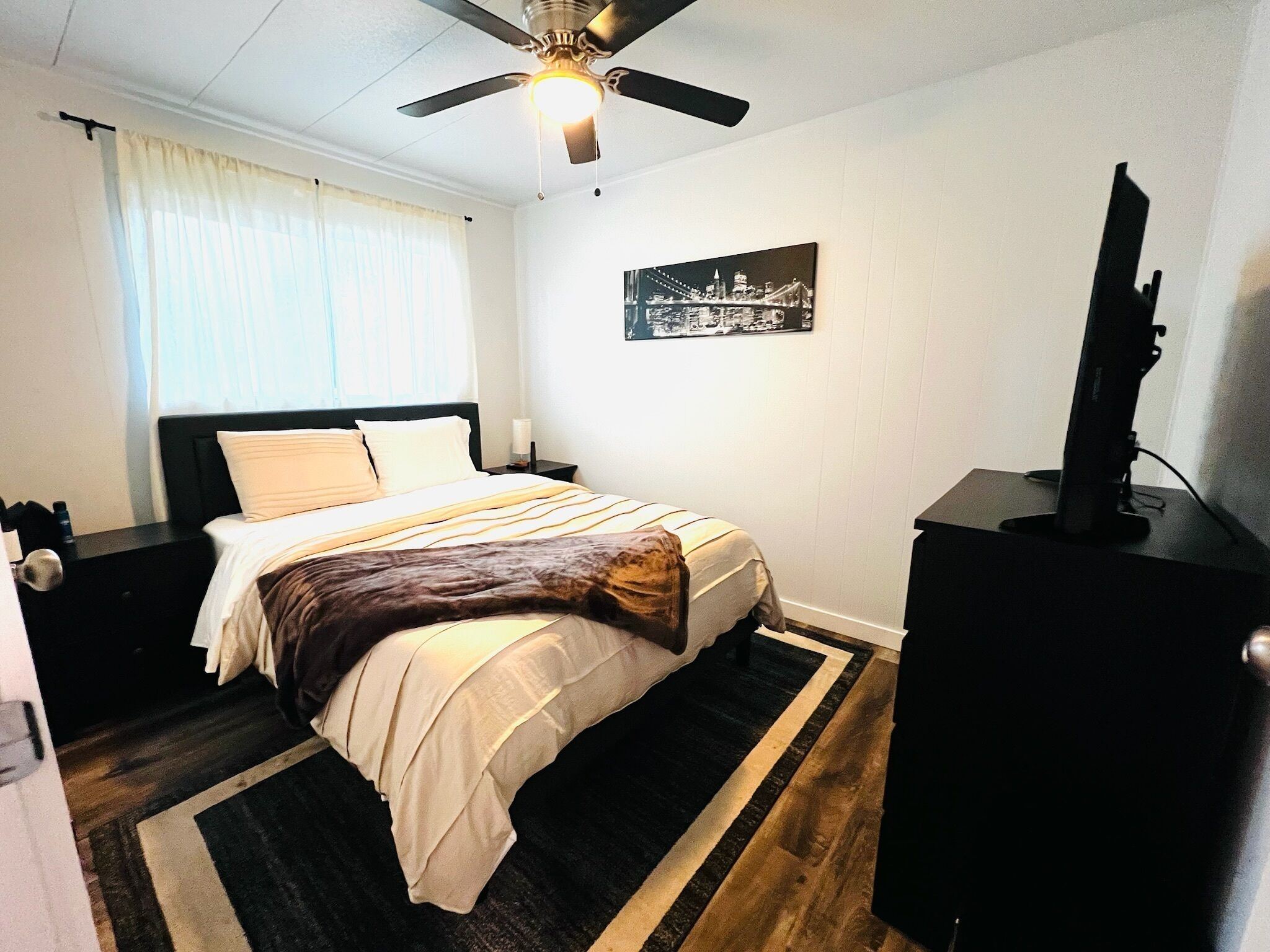 A hotel room with a bed, a ceiling fan, a nightstand, a TV, and a cityscape painting on the wall.