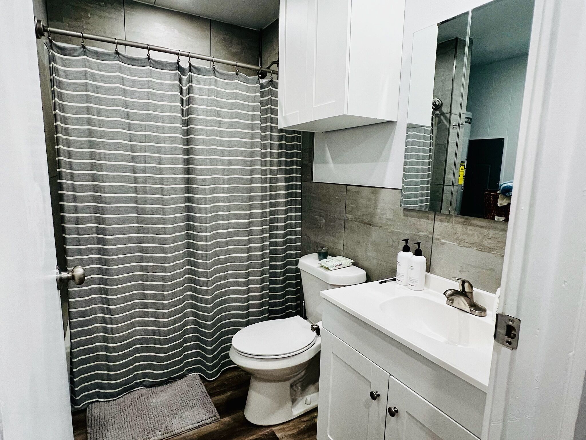 A bathroom with a striped shower curtain, a white toilet, a sink with a mirror above it, and a small rug on the floor.