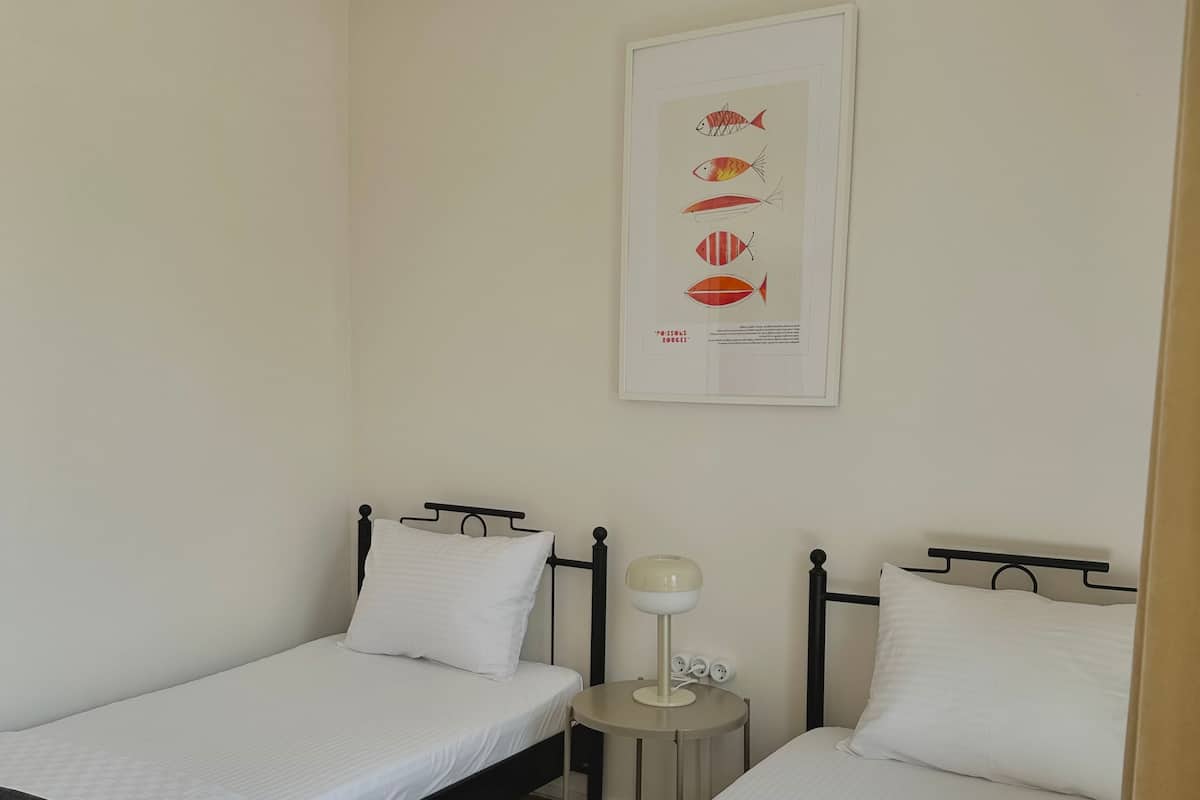 A hotel room with two single beds, a small bedside table with a lamp, a framed fish illustration on the wall, and a window with a view of the outside.